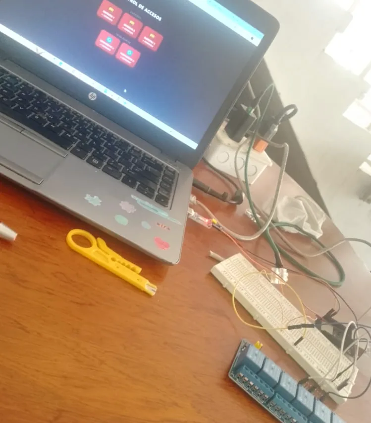 Laptop mostrando la interfaz de control de accesos con los botones de cada barrera y torniquete, junto a un protoboard y herramientas de red.